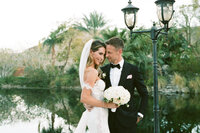 Bride and groom embracing outdoors by water, bride holding bouquet