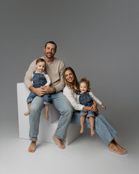 Family of four sitting together on white studio cubes with soft gray background, coordinated neutral outfits captured by Kaitlyn Dawn Photography, studio family photographer in Inland Empire.