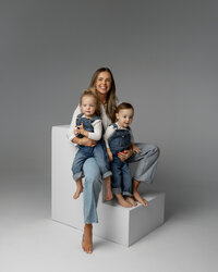 Mother sitting on white studio cubes holding her two young children against a soft gray backdrop, timeless studio family portrait by Kaitlyn Dawn Photography, Yucaipa family photography studio.