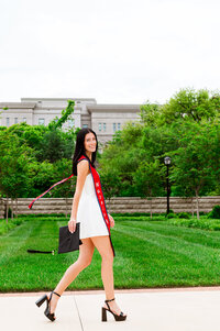 College senior wearing white dress and walking toward the Medical College at Belmont, holding her cap with red stole floating in the wind