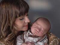 Mother with fringed hair cradling a sleeping newborn wrapped in white, photographed in warm indoor light.