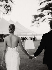Couple holding hands in black and white as they approach a softly lit reception tent at twilight during their waterfront wedding.