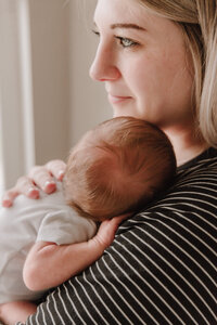 A mom faces a window with her newborn son sleeping against her shoulder.
