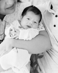 Black and white close-up of an awake newborn baby looking toward the camera while resting in mom and dad’s arms during an in-home session.