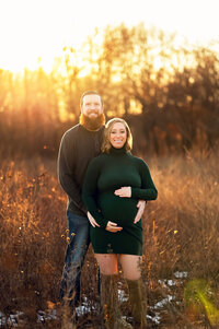 Expecting couple standing in a sunlit Wisconsin field during golden hour, with the pregnant woman wearing a forest green dress and her partner embracing her from behind.