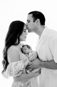 Dad kissing mom while she holds her newborn baby during Miami newborn photoshoot