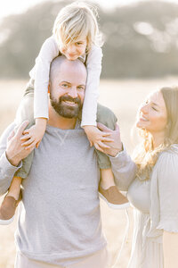 Close up family photo of a mother looking at her son as he is sitting on his dads shoulders.