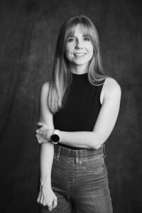 A black and white picture of a blonde woman with straight hair smiling in front of a dark gray backdrop. She wears jeans and a black tank top. She rests one arm by her side, holding it with her opposite hand.