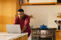 Man at a laptop on the kitchen counter, taking a call while cooking