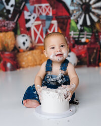 Smiling baby in denim overalls with cake-covered hands during cake smash. Photograph by Yucaipa cake smash photographer Kaitlyn Dawn Photography.