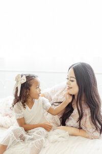 A light filled motherhood photo in a photography studio of a mother laying on a bed as her daughter gently holds her face as they smile at each other.