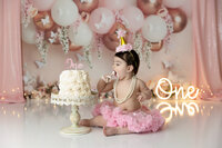 Fist birthday girl wearing a pink tutu and gold party hat eating cake during her Cake smash photo session.