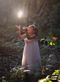 Young girl in a soft pink dress gazes in wonder at a glowing fairy hovering above her hands during a fairy adventure signature in St. Johns. - A portrait experience by Nature's Child Photography.