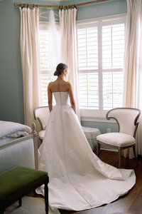 Bride standing in front of a sunlit window in her strapless wedding gown, looking outside before the ceremony at Burritt on the Mountain in Huntsville, Alabama, photographed by Perry Hancock.