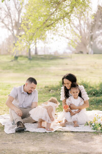 Family of four enjoying a candid outdoor portrait session in a sunlit park, sitting on a blanket as parents smile with their young children during a relaxed lifestyle photography moment.