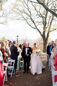 Bride walking arm-in-arm with her father down the aisle during the wedding ceremony at Burritt on the Mountain in Huntsville, Alabama, as guests look on, photographed by Perry Hancock.