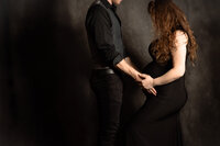 A close up picture of a pregnant woman with her husband on a gray backdrop. She holds his hands to her belly and holds one of her legs slightly forward in the frame. Her long brown hair covers her face, and her husband's face is just out of frame.