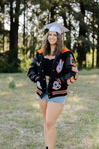 High school graduate wearing a cap and letterman jacket standing in a grassy field, graduation portrait captured by Kaitlyn Dawn Photography.