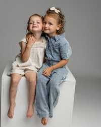 Two young siblings sitting together on a white studio cube, smiling and hugging in a neutral light gray studio setting, photographed by Kaitlyn Dawn Photography, timeless studio family photographer specializing in natural children’s portraits.