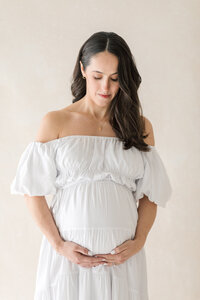 A woman wearing an off the shoulder white dress cradles her pregnant belly while gazing down at it.