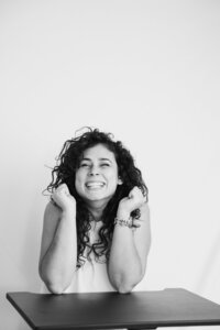 A black and white picture of a woman with dark curly hair smiling joyfully as she sits in front of a white backdrop. She wears a white dress and rests her elbows on a black table while lifting both fists with excitement.