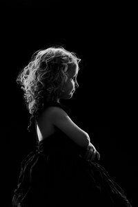 A black and white picture of a young blonde girl sitting upright against a black background. The lighting is dramatic, creating a silhouette around her.