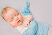 Peaceful newborn baby boy wrapped in blue knit against cream background