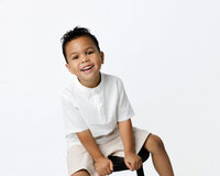 Young boy sitting on a stool in a neutral studio with soft lighting, wearing a white shirt and smiling during a modern child portrait session by Kaitlyn Dawn Photography.