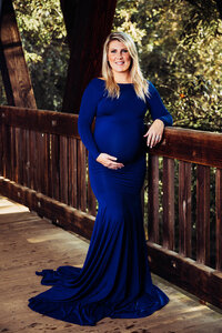 A pregnant woman in a royal blue dress leans against the railing on a covered wooden bridge. She smiles with one hand propped under her belly.