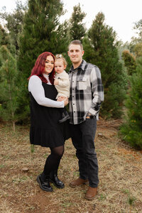 Parents standing together with their baby in a wooded outdoor setting, coordinated neutral outfits captured by Kaitlyn Dawn Photography, lifestyle family portrait photographer.