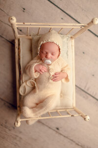 Cozy baby photo of a newborn in a cream knit romper and bonnet, sleeping in a vintage bed at a Brookfield-area newborn photo studio.