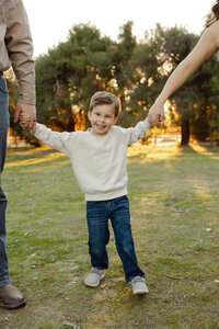Young boy smiling while holding his parents’ hands in a grassy park at sunset, playful lifestyle family photography by Kaitlyn Dawn Photography.