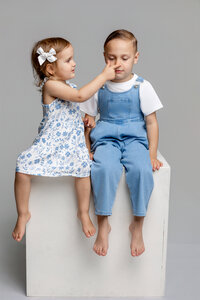 Brother and sister sitting together on a white studio cube in a minimalist gray studio, playful and candid sibling portrait by Kaitlyn Dawn Photography, professional family photography studio.