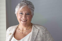 Smiling woman with short silver hair in a cream tweed jacket and gold necklace, photographed in bright natural light.Person in a black leather jacket and sunglasses caught mid-jump on an airfield tarmac, propeller aircraft behind.