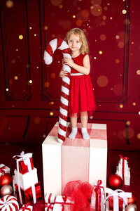 A young blonde girl in a red dress stands on a large white box wrapped with red ribbon and surrounded by red and white Christmas decorations. She holds a large candy cane decoration and smiles sweetly.