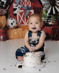 Smiling toddler with cake on his face and hands, celebrating his first birthday in a farm-style studio setup. Photograph by Yucaipa cake smash photographer Kaitlyn Dawn Photography.