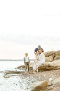 Beautiful lake side family photo as the parents are by the rocks hugging their daughter as their son plays by the water.