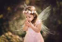 Little girl in a blush pink fairy dress and floral crown cups her hands and blows sparkling fairy dust with delicate wings during a fairy adventure signature in O’Fallon, Illinois. - A portrait experience by Nature's Child Photography.