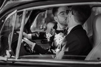 Mariés s'embrassant à bord d'une voiture d'époque, boutonnières florales et nœuds papillon, portrait intime en noir et blanc près du Pont Couvert