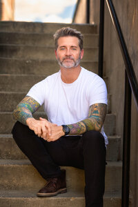 A bearded man with sleeve tattoos sits on a set of concrete steps. He smiles softly.