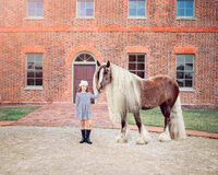 Young girl in white lace dress standing beside a white horse in soft golden light