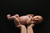 A naked baby lies in her father's hands against a black backdrop.
