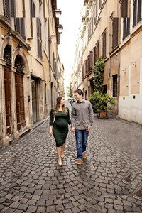 Photo of pregnant woman in a green dress holding her husbands hand while looking at each other on a classic Roman street. Taken by Rome Photographer, Tricia Anne Photography