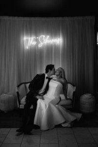 bride and groom sitting in a chair below a light up sign with their last name