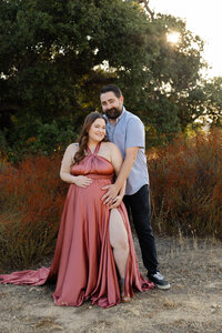 Romantic outdoor maternity portrait of expecting couple embracing with father holding mother's baby bump while she wears a flowing terracotta maternity gown during a sunset lifestyle maternity photoshoot by Kaitlyn Dawn Photography.