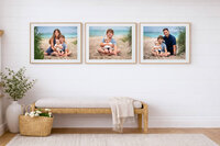 Three framed beach family portraits displayed above a woven bench in a light coastal entryway in Eastern North Carolina