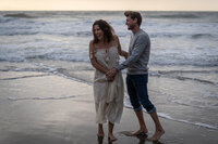 beautiful couple holding hands and laughing as they walk down the beach at sunset without shoes