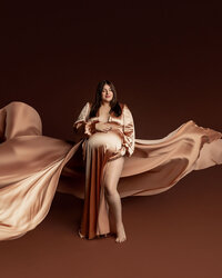 Pregnant woman wearing a flowing champagne satin maternity gown with dramatic fabric movement during a luxury studio maternity photoshoot, photographed by Kaitlyn Dawn Photography.