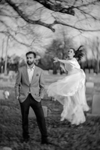 Black and white image of a man in a light suit standing among gravestones as a woman in a white gown moves behind him.