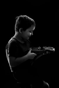 A black and white picture of a boy holding a model car. He smiles at it as the dramatic lighting creates a subtle silhouette around him.